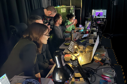 People working in at a desk in front of computer monitors and laptops in a dimly lit studio.