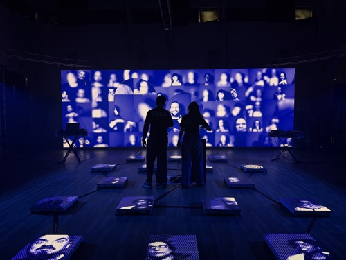 Two people standing in front of a virtual production wall displaying an installation of faces, in a dark studio.